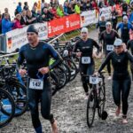 Triathleten in Neoprenanzügen laufen am frühen Morgen zu ihren Rädern in der Wechselzone eines Triathlon-Wettkampfs an einem See in Deutschland.