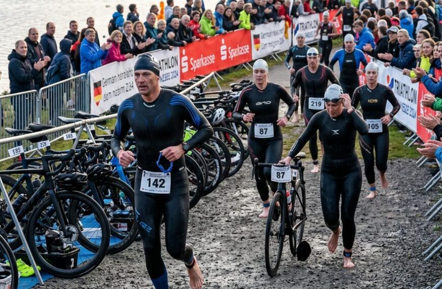 Triathleten in Neoprenanzügen laufen am frühen Morgen zu ihren Rädern in der Wechselzone eines Triathlon-Wettkampfs an einem See in Deutschland.