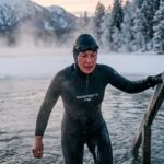 Winterschwimmer in Neopren-Kappe und Schwimmbrille steigt in einen eisigen See, Wasserdampf steigt auf, schneebedeckte Bäume im Hintergrund